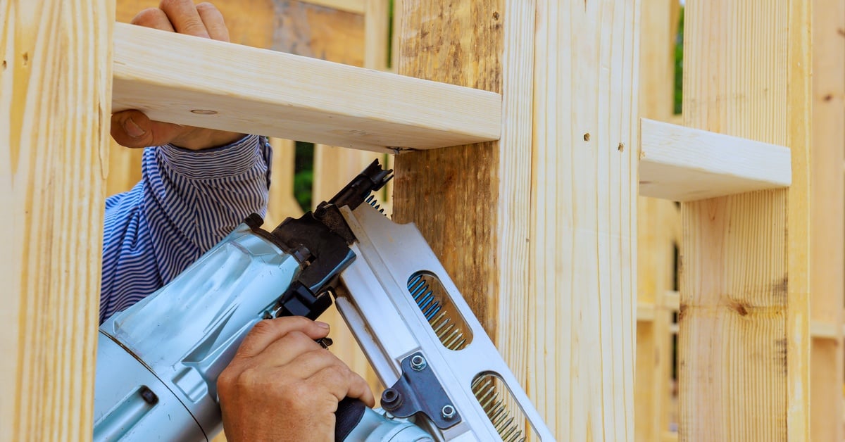 HA person's hand holds a heavy-duty professional nail gun, nailing the wooden frame structure of a house.