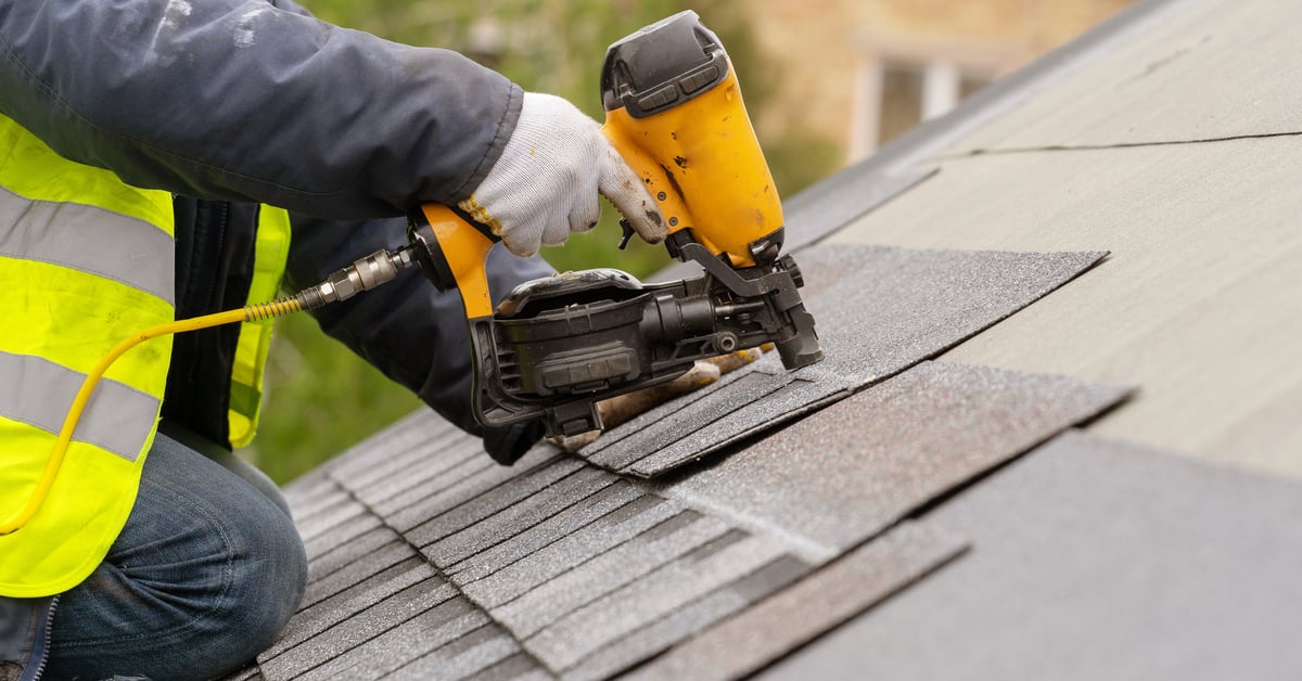 A construction worker kneels on a gray roof using a pneumatic nail gun to staple a tile. They wear gloves while using the tool.