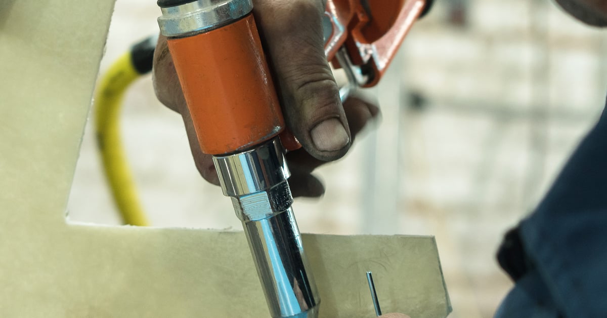 A male mechanic's hand is holding a pneumatic riveting tool. The tool is orange and has a stainless steel head.