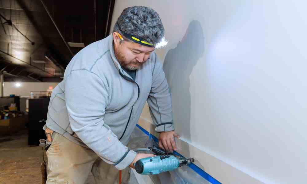 A male contractor wearing khaki pants and using an air-powered nail gun to install wood trim on a wall inside of an office.