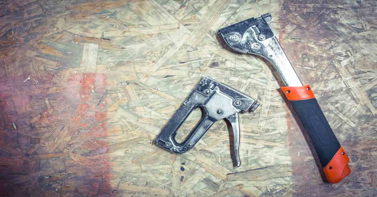 A couple of staplers on wood. The one on the left is shorter, while the one on the right has a long handle.