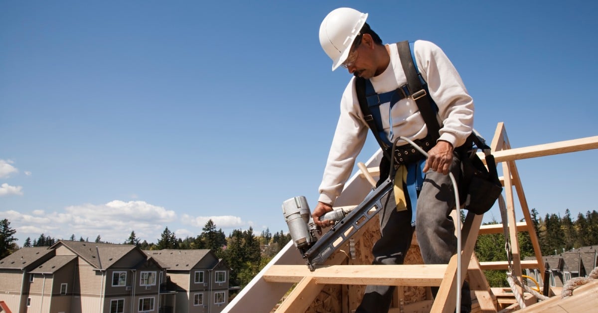 A worker wearing a white hard hat and safety harness using a framing nailer on the roof of a building.