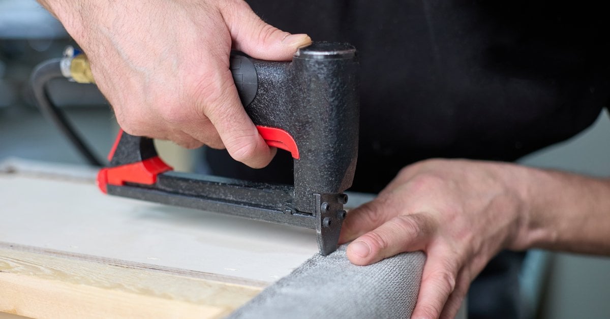 A man is using a small pneumatic staple gun to insert staples. He is fixing upholstery fabric to wood.