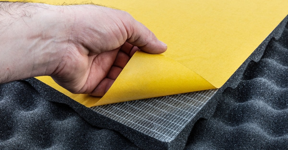 Close-up of acoustic foam with a hand peeling off the yellow underside, showing adhesive coating underneath.
