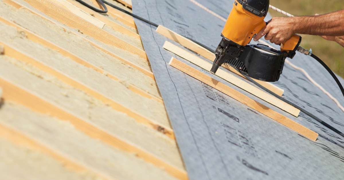 A contractor holding a pneumatic nail gun on a roof. The surface has exposed wood boards and a gray underlayment.