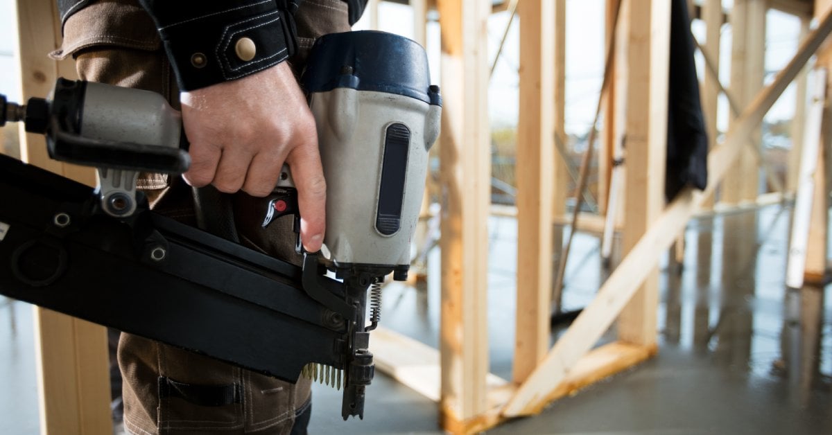 A man wearing brown pants and a blue shirt stands in an unfinished home while holding a finish nailer nailgun.