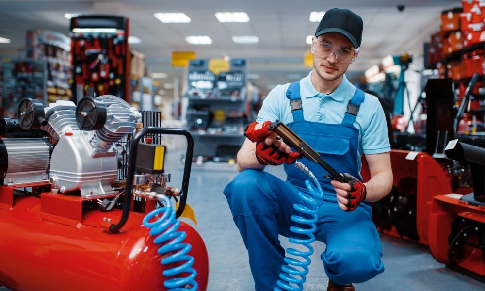 A technician in a workshop handles a pneumatic tool with safety gear. He is inspecting equipment near an air compressor.
