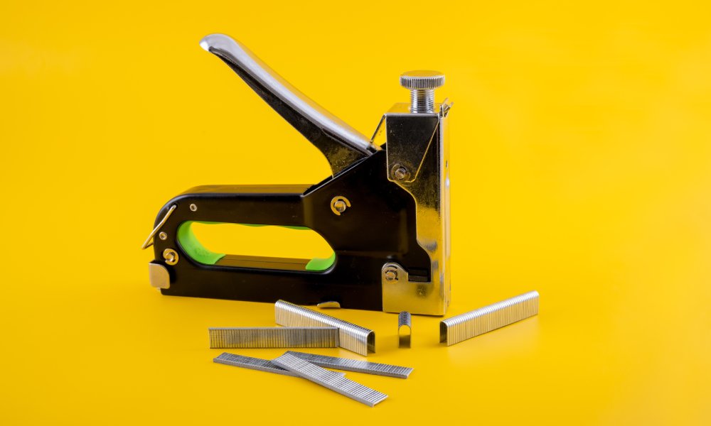A manual industrial stapler sits in the middle surrounded by staples of different sizes on a yellow background.