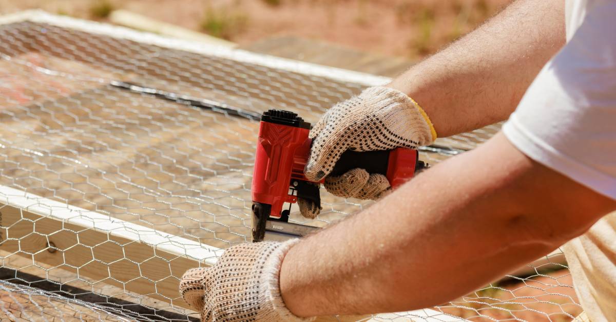 A person wearing gloves secures chicken wire to a wooden frame using a red hog ring stapler on a sunny outdoor workspace.