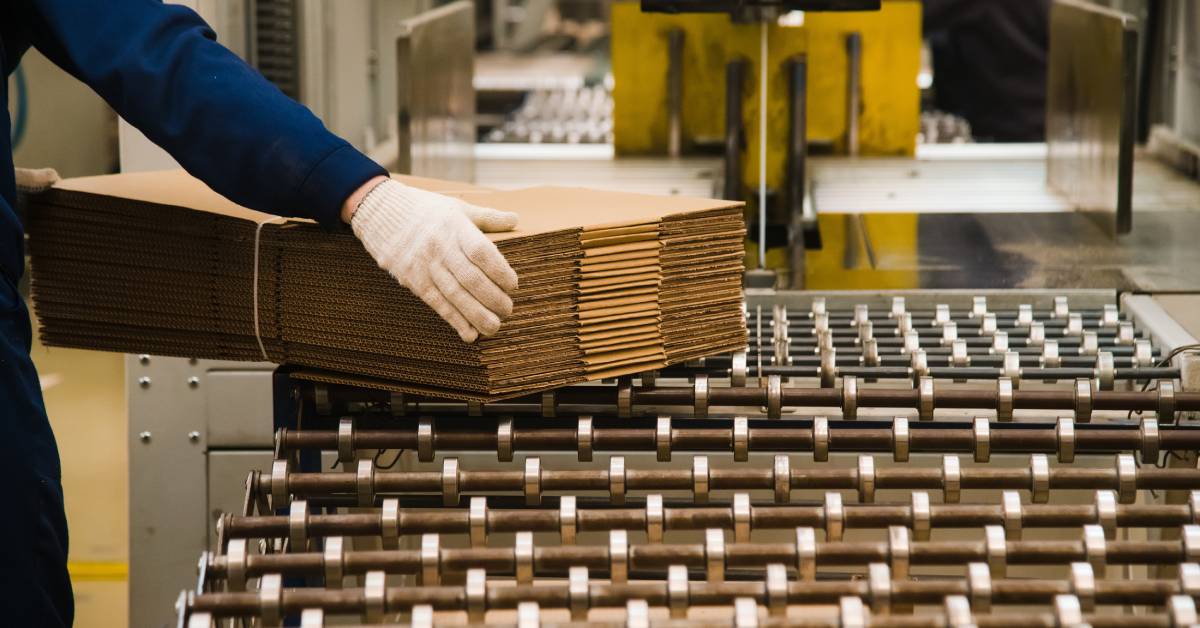 A person wearing blue long sleeves holds a stack of corrugated cardboard sitting on top of a production line.