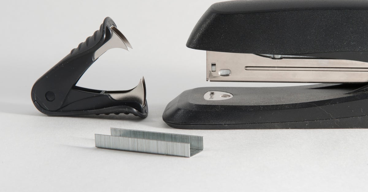 A black stapler and stapler remover face each other, with a staple refill in front. All three pieces are on a white background.