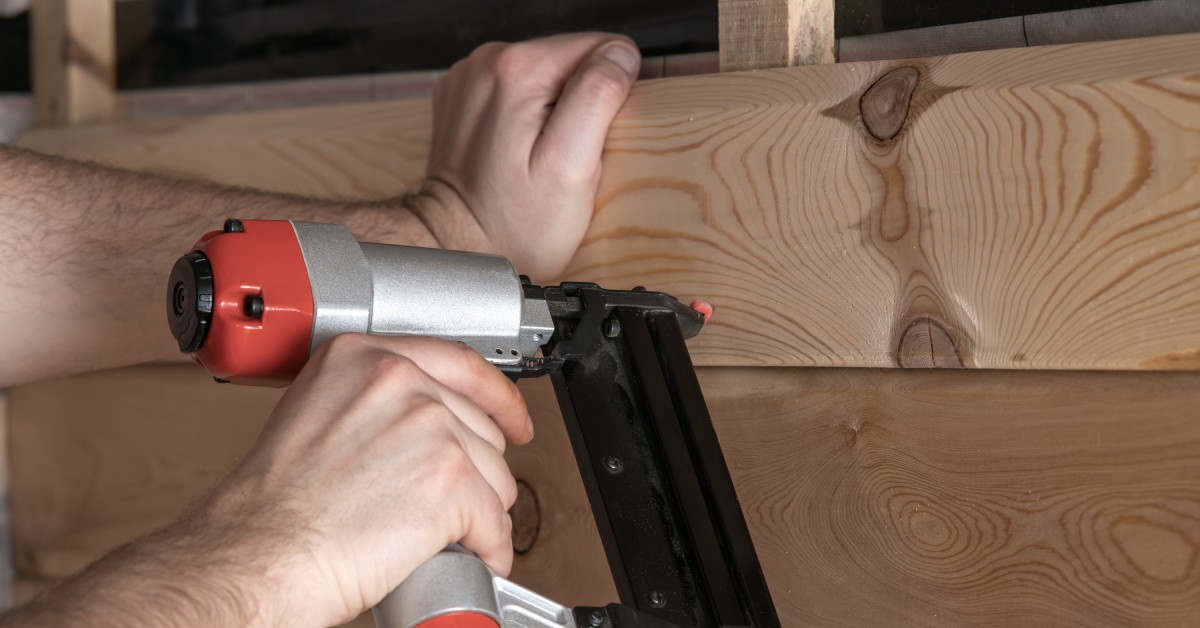 A person holds a silver and red pneumatic stapler while pushing at a wooden panel with the other hand.