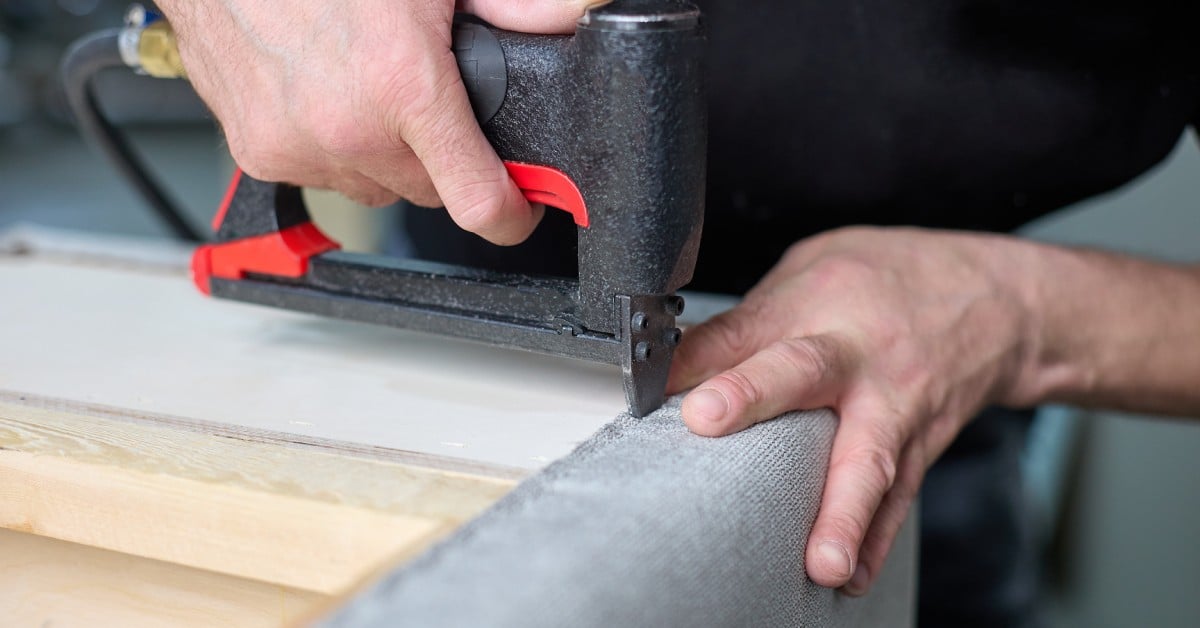 A person wearing a black shirt uses a pneumatic staple gun to staple the fabric on a piece of furniture.