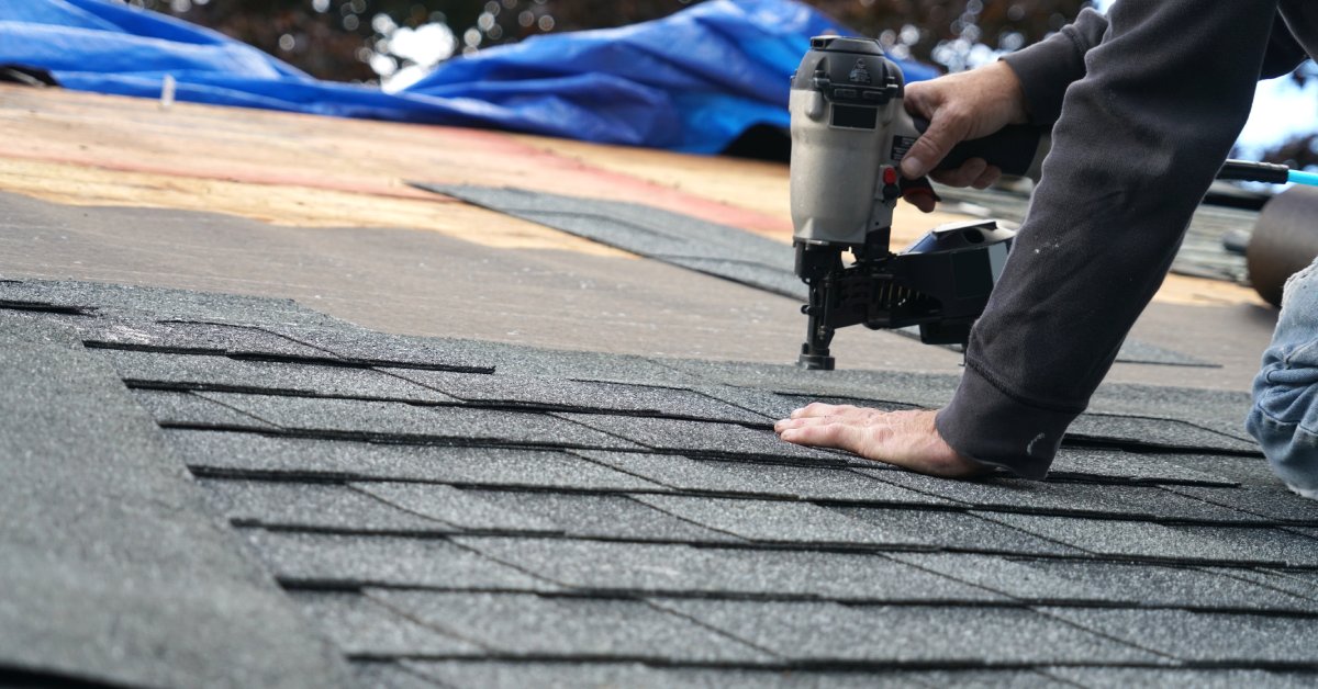A worker in a hoodie using a nail gun to install black asphalt shingles on a roof, with a blue tarp in view.