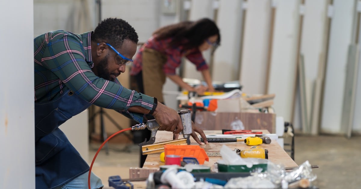A male carpenter wearing protective glasses uses a heavy duty staple gun to make furniture in a production line.