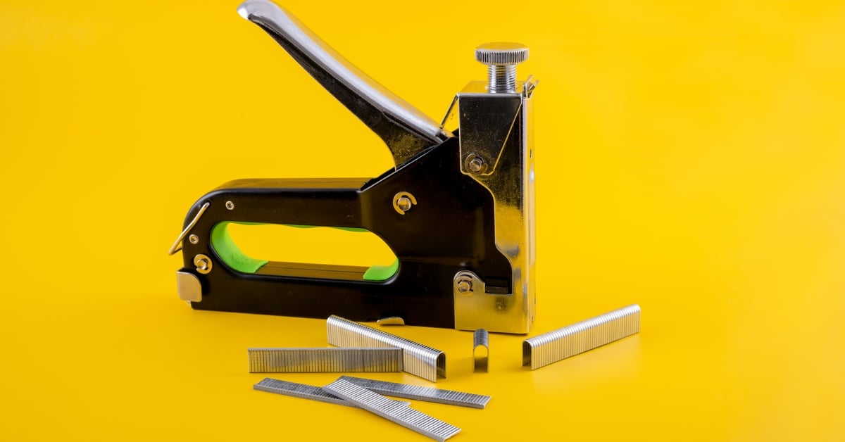 A mechanical stapler sits next to different types of fasteners, including brads, against a bright yellow background.
