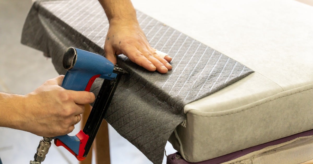 Close-up of a worker’s hand operating a blue pneumatic stapler in a workshop for upholstery and assembly work.