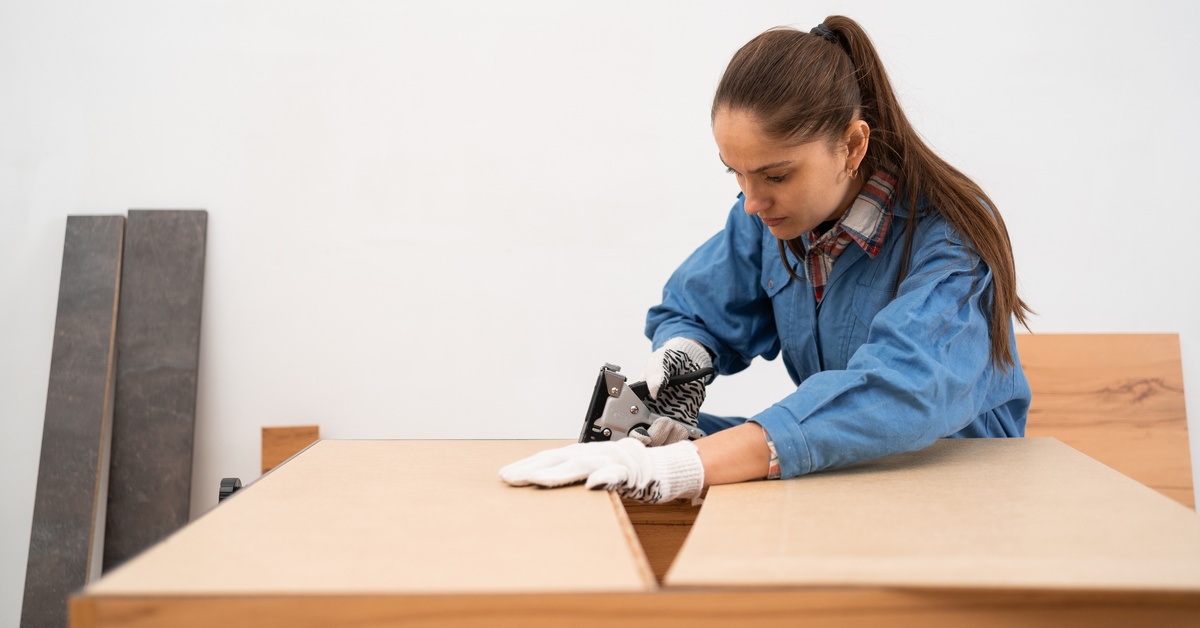 Carpenter assembling furniture uses a stapler to attach fabric to a wooden frame on a workbench in a workshop.