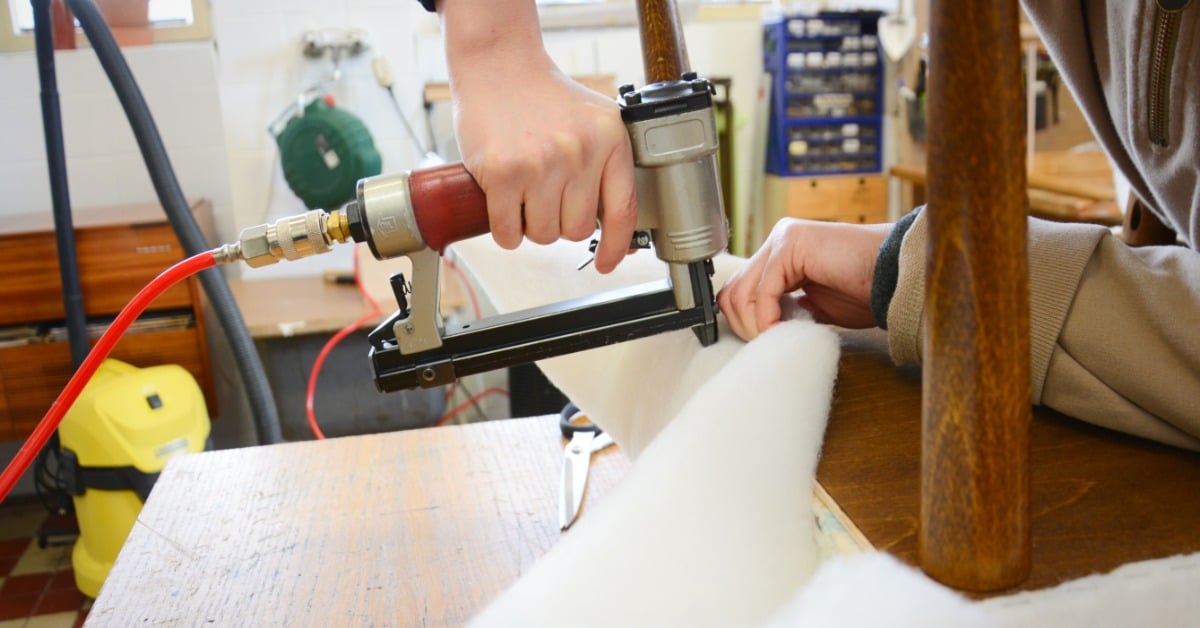Close-up of a woman&rsquo;s hands using a pneumatic stapler on fabric and padding in an upholstery workshop.