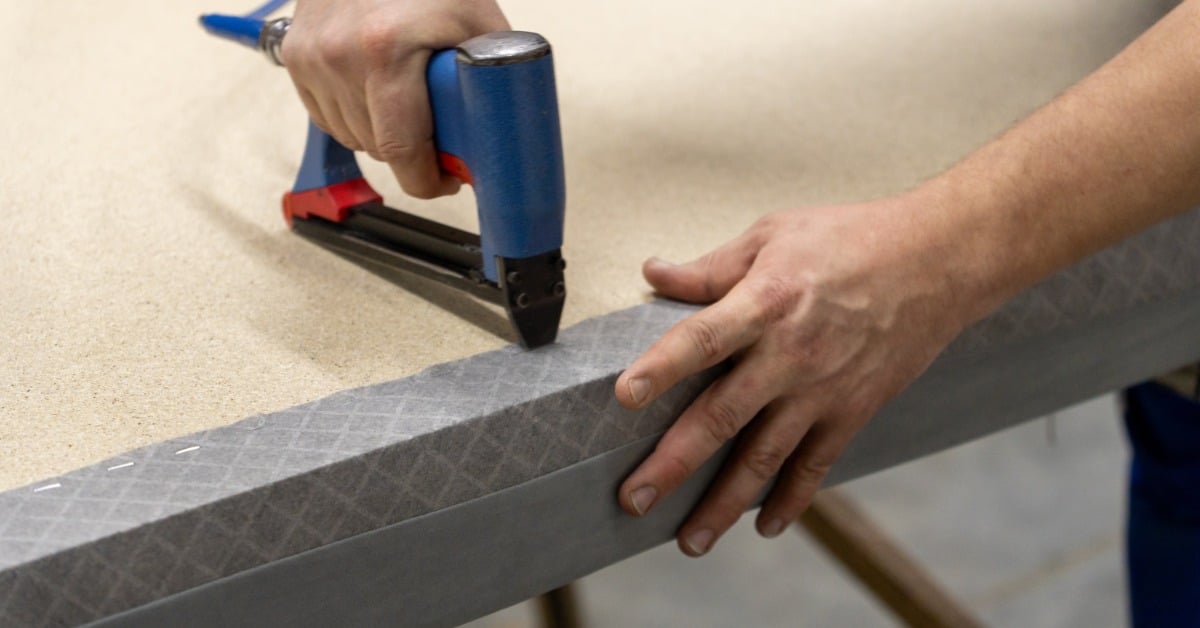 A person using a pneumatic stapler on light gray colored fabric while working on upholstery in a workshop.