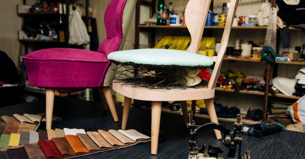 Two chairs, fabric swatches, tools, and supplies arranged on a worktable inside an upholstery workshop.
