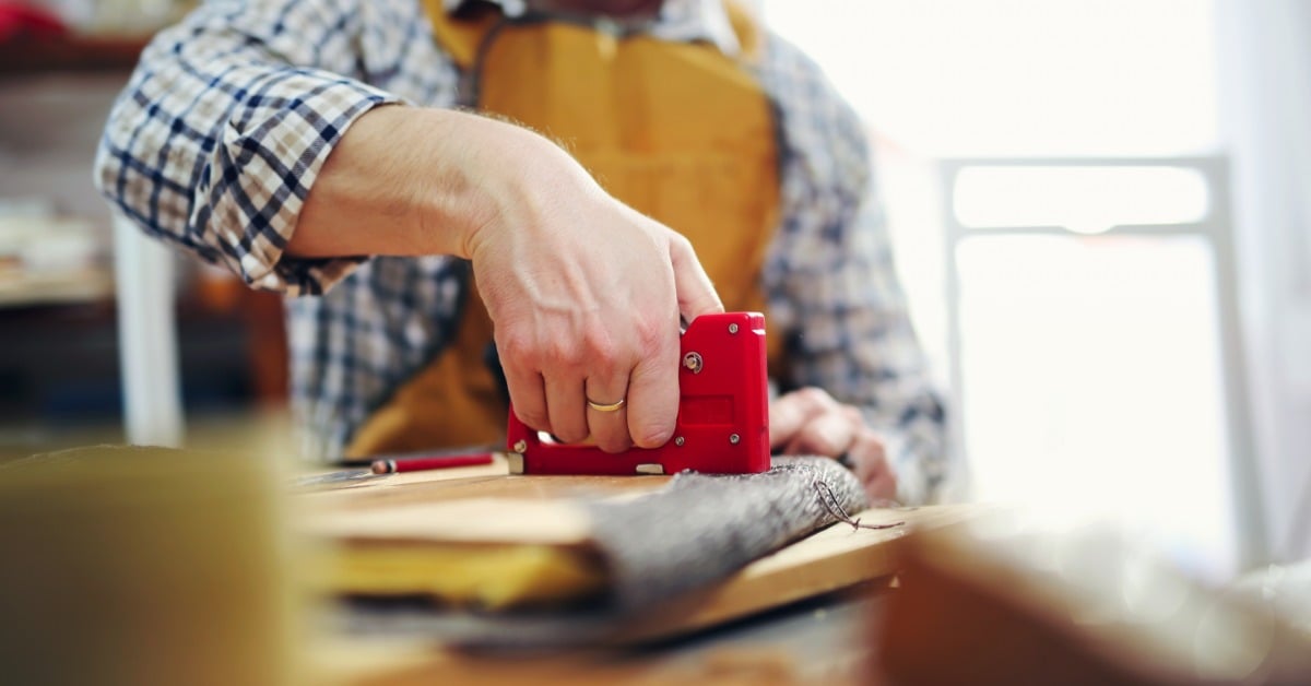 A person wearing an apron using a red staple gun on fabric laid across a wooden workshop table near a bright window.