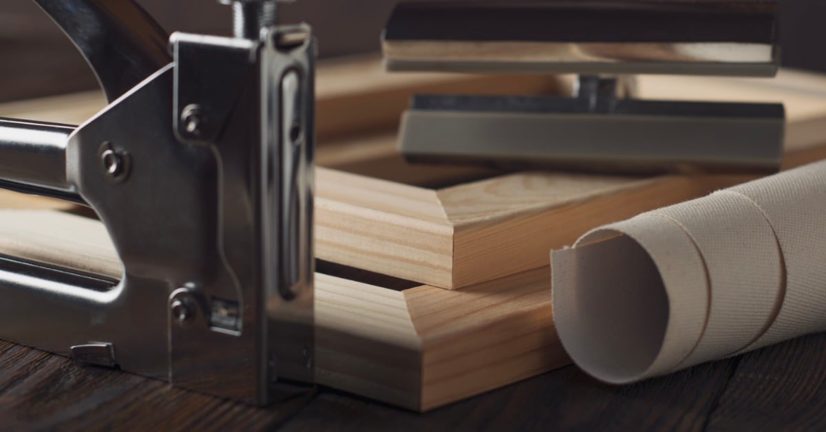 a rolled canvas and stapler beside wooden stretcher bars and a tensioning tool on a light tabletop in soft focus.