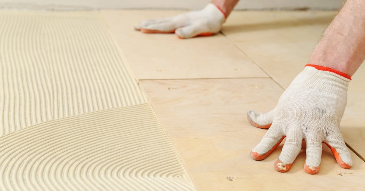A person in white work gloves presses plywood sheets beside ridged adhesive spread across a floor during installation.