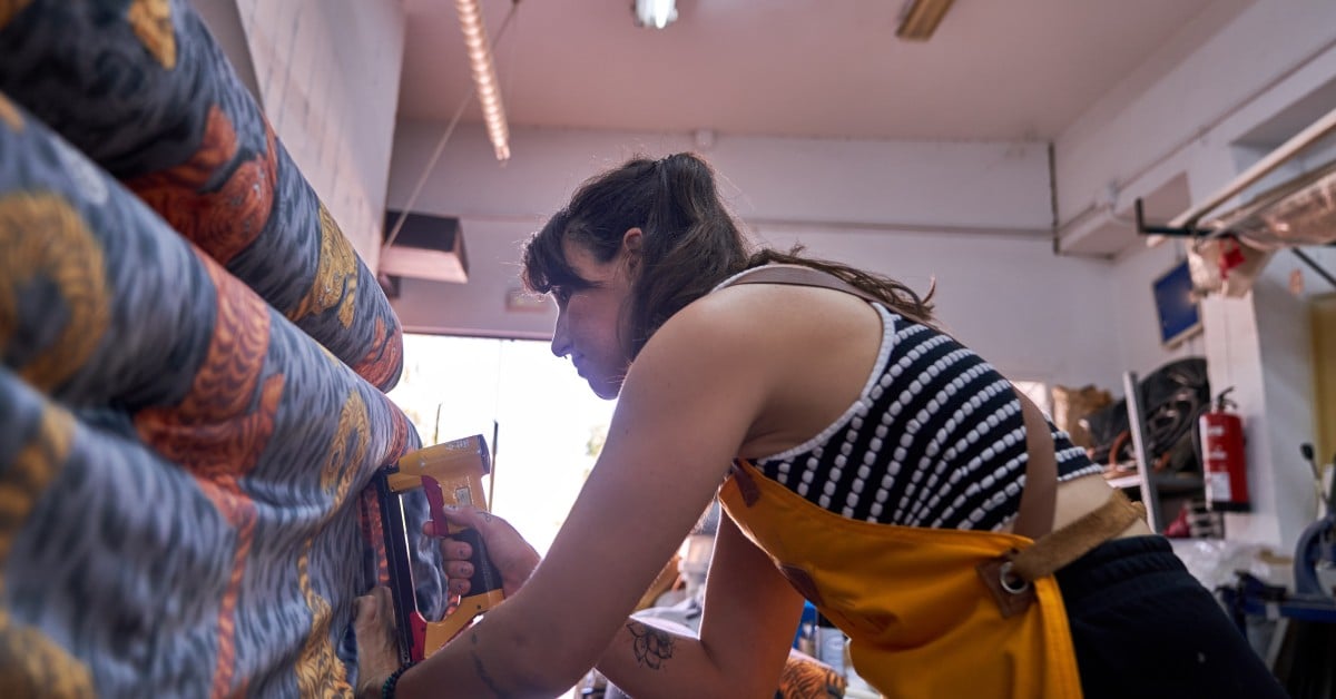A female upholsterer uses a yellow staple gun to apply colorful fabric to the furniture in a workshop.