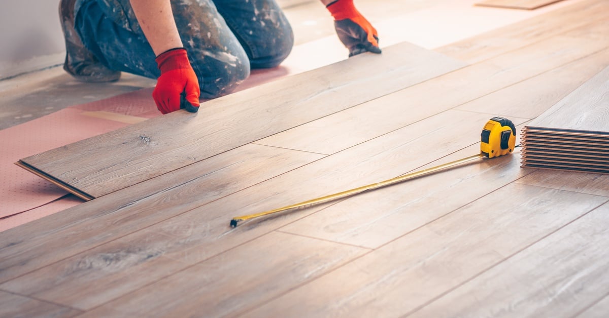 A person wearing jeans and red gloves kneels and lays down laminated wood flooring. On top of the planks is a measuring tape.