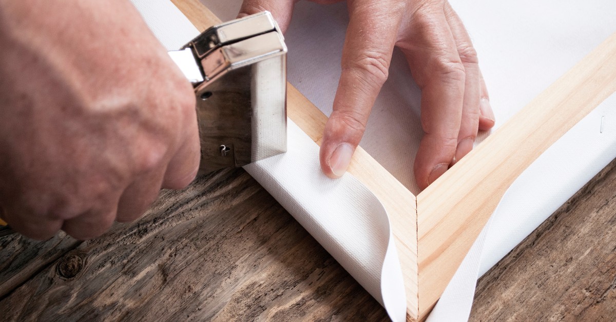 Close-up of a man's hands as he uses a framing stapler to attach a white canvas to a wooden picture frame.