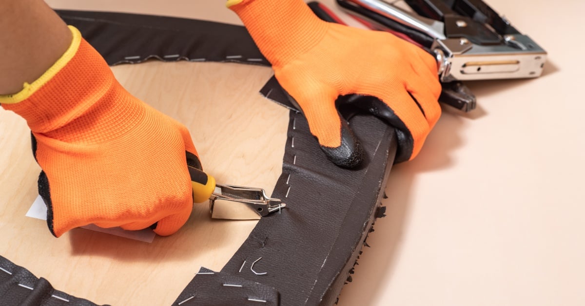 A person wearing orange gloves uses an upholstery staple remover to remove the staples on a wooden chair.
