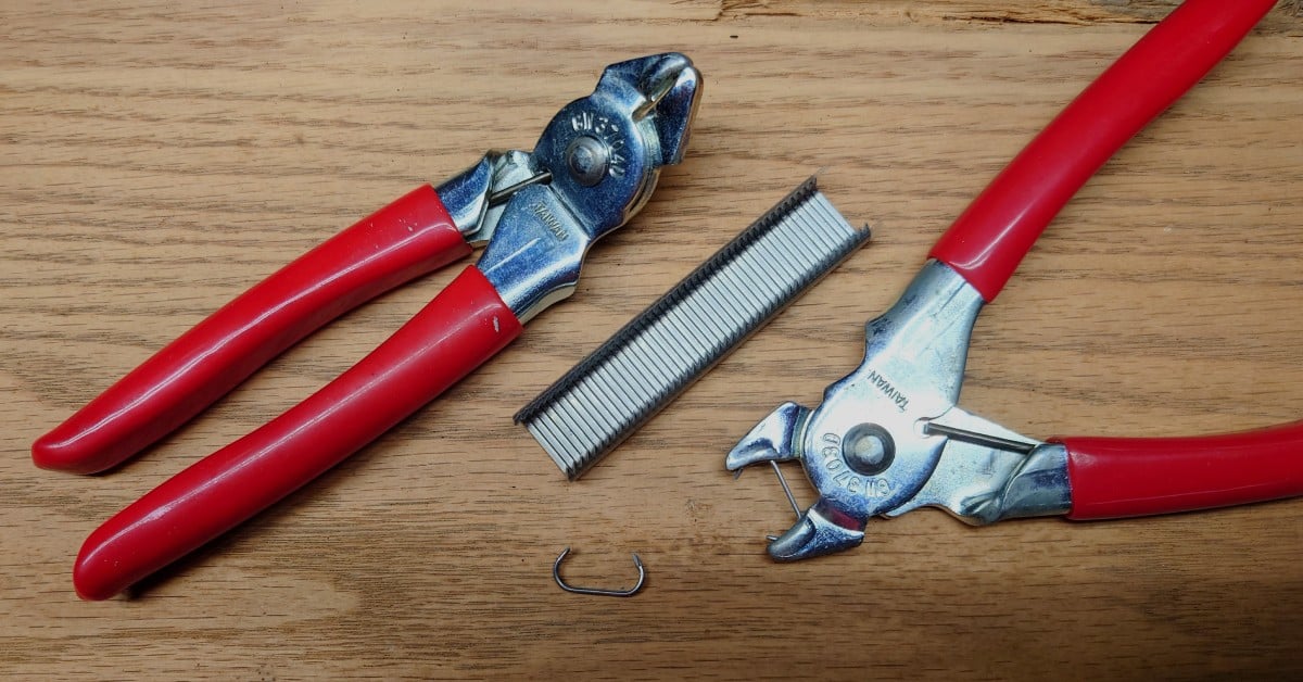 Two hog ring pliers sit next to each other on top of a wooden table, with staples in between them.