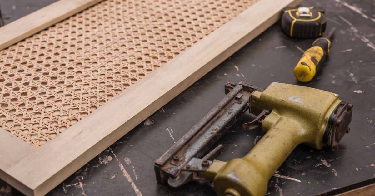 A wooden rattan weave door sits on top of a black table, next to a measuring tape and yellow staple gun.