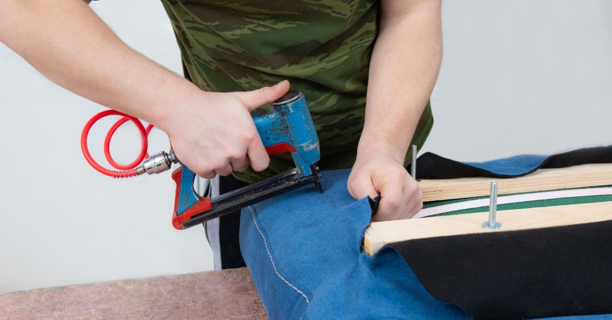 A person wearing a green camo shirt uses a blue pneumatic staple gun to attach the blue fabric onto furniture.