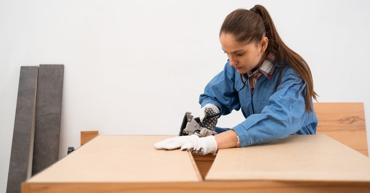 Carpenter assembling furniture uses a stapler to attach fabric to a wooden frame on a workbench in a workshop.