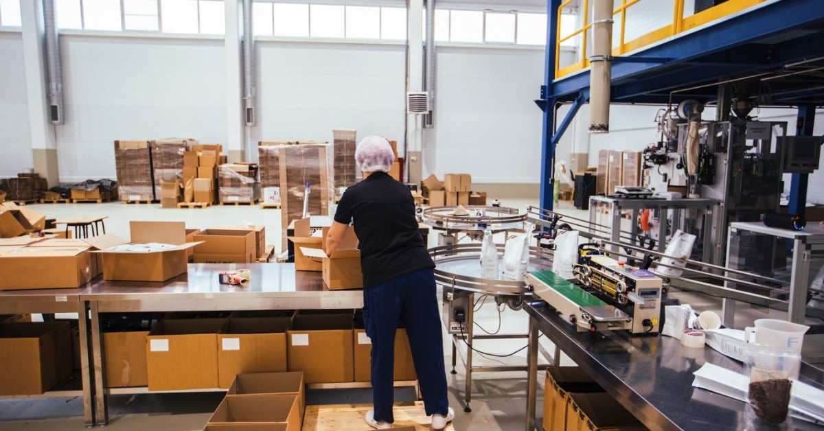 A worker packs sealed coffee bags into open cardboard boxes on a table in a warehouse or packing area.