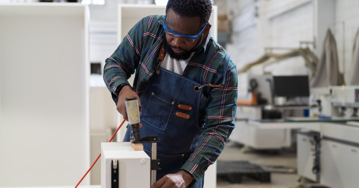 Carpenter uses a pneumatic staple gun to fasten a white wood plank on a workbench inside a wood workshop.