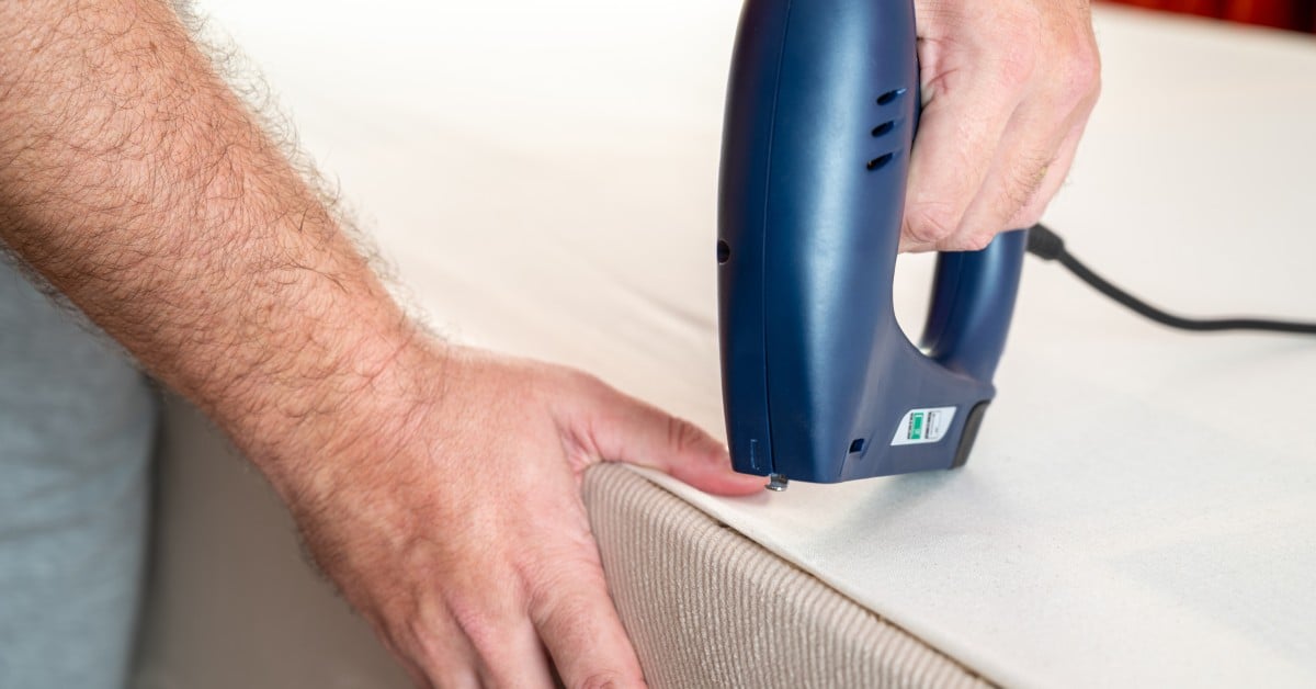 A person touches a beige sofa with one hand and uses a blue electric staple gun with the other hand.