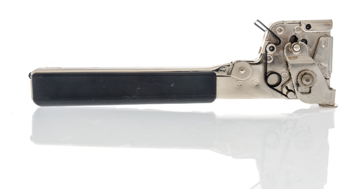 A silver hammer tacker with a black handle lays flat on a white, glassy surface while it reflects a shadow.