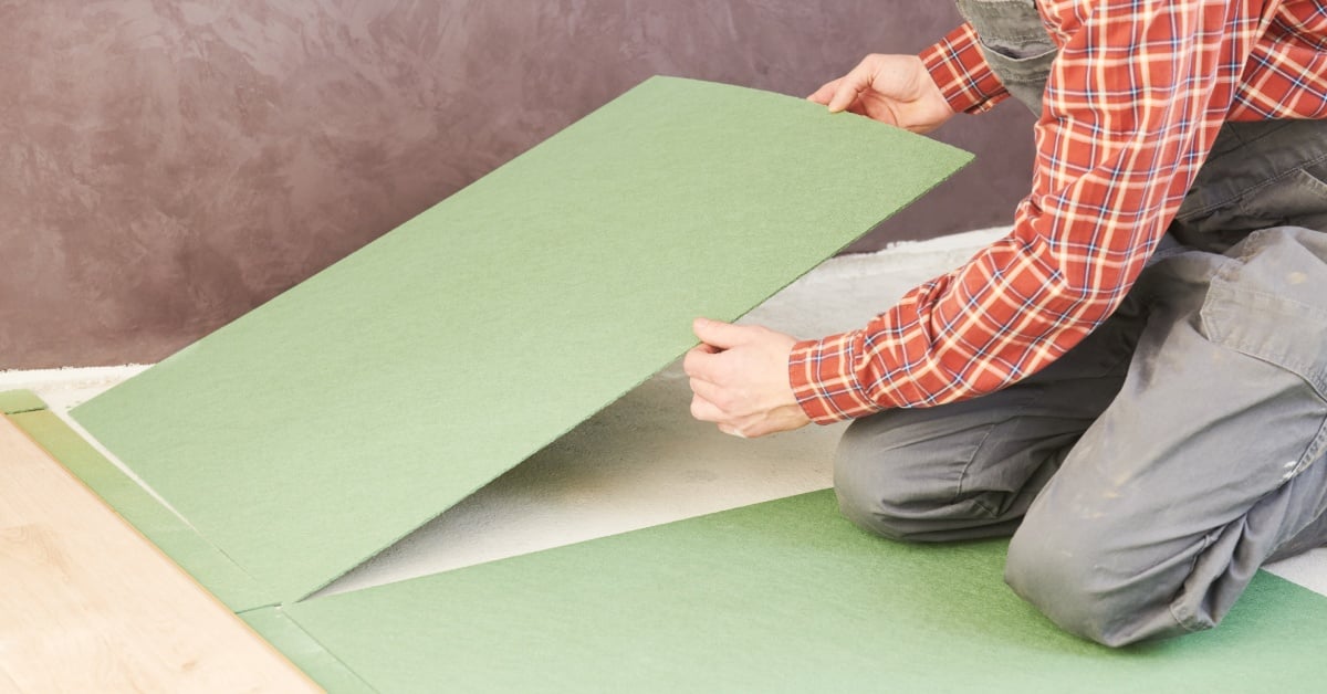 A person wearing a red plaid shirt and gray pants kneels on the floor and lays down a green underlayer padding.