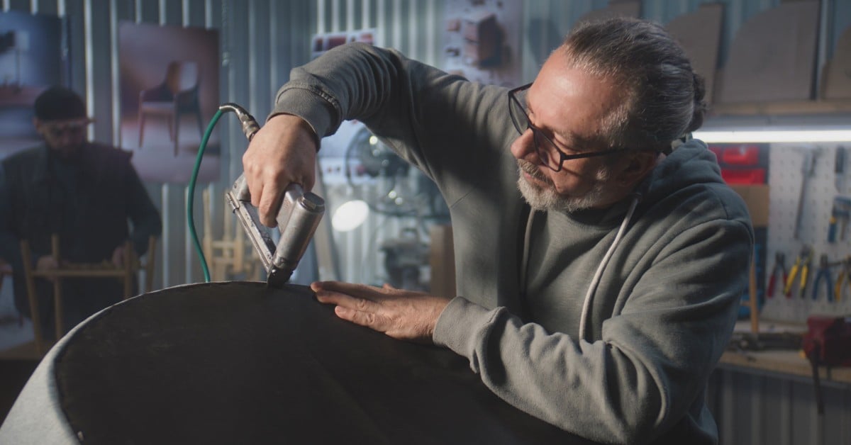An older male wearing glasses and a gray sweater uses a staple gun to attach the fabric on a piece of furniture.