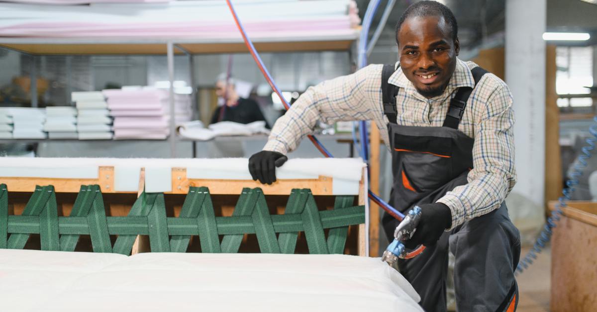 A male furniture maker wearing black overalls smiles while using a pneumatic staple gun as he assembles a sofa.