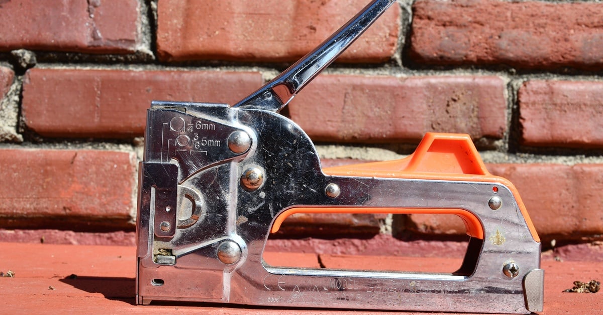 A metal and orange staple gun with a raised handle resting on a red brick ledge against a brick wall.
