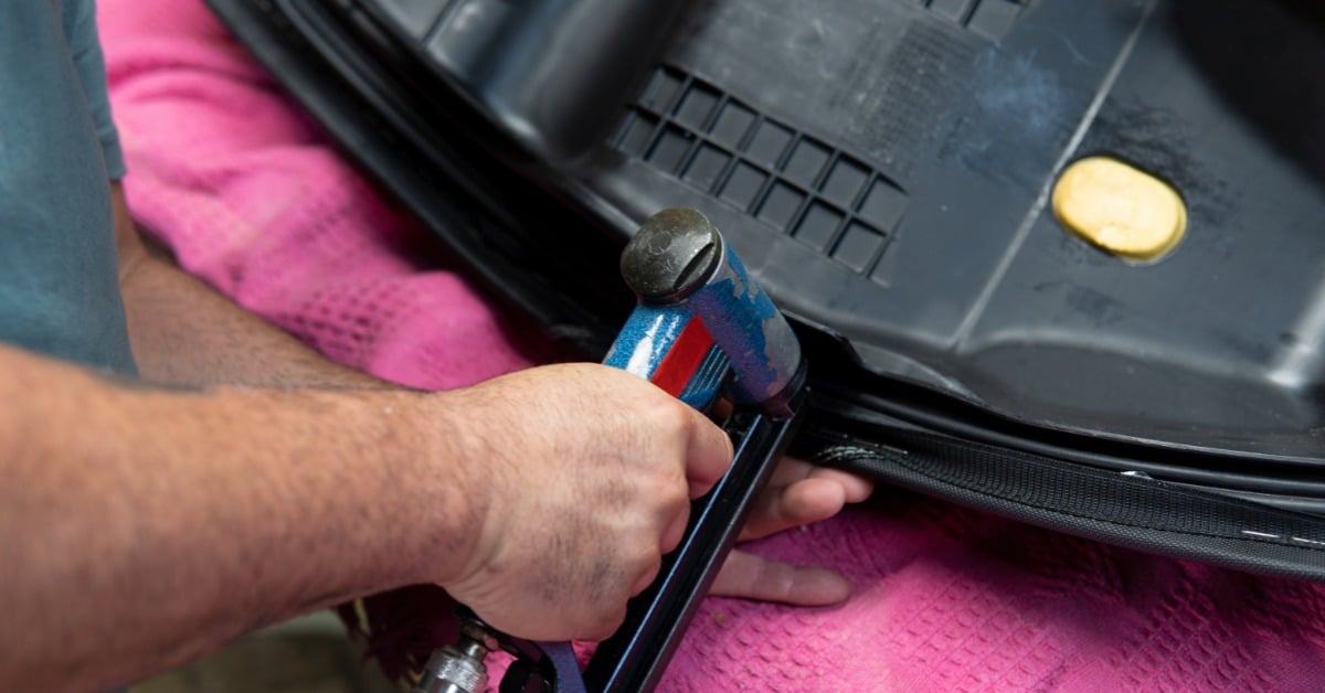 A person's hands using a pneumatic stapler on the underside of a black seat resting on a pink towel.