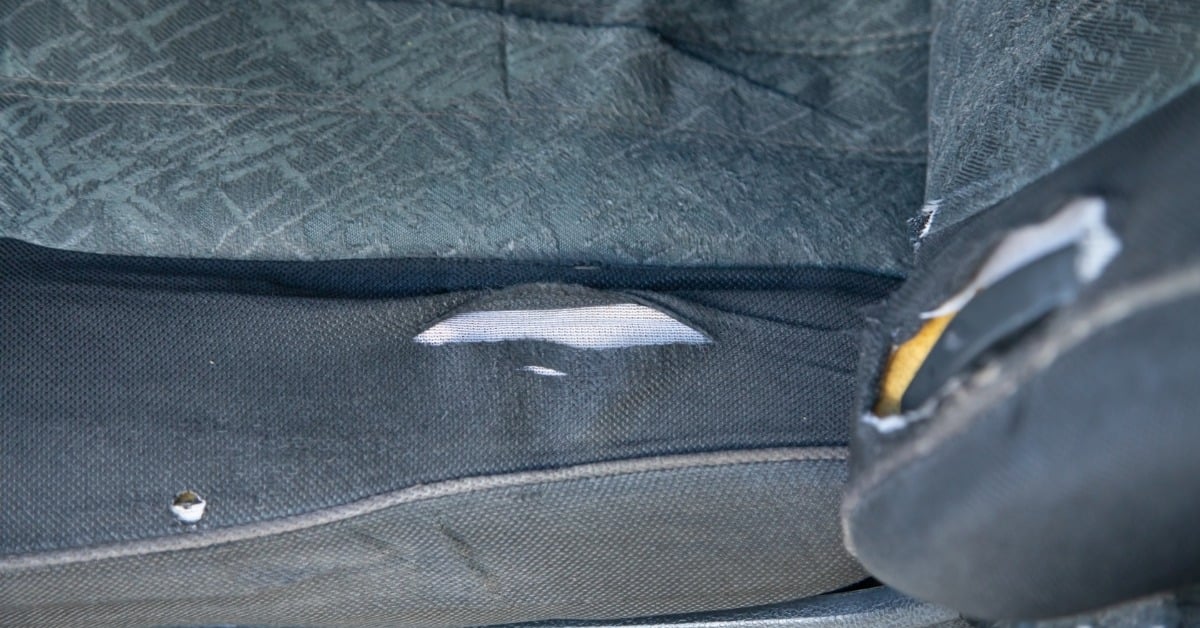 A worn dark fabric car seat with several torn areas, frayed edges, and white padding visible through the upholstery.