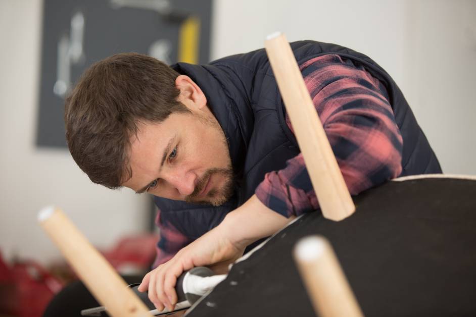 A male carpenter works on reupholstering a stool by turning it over to attach new fabric with a tool.